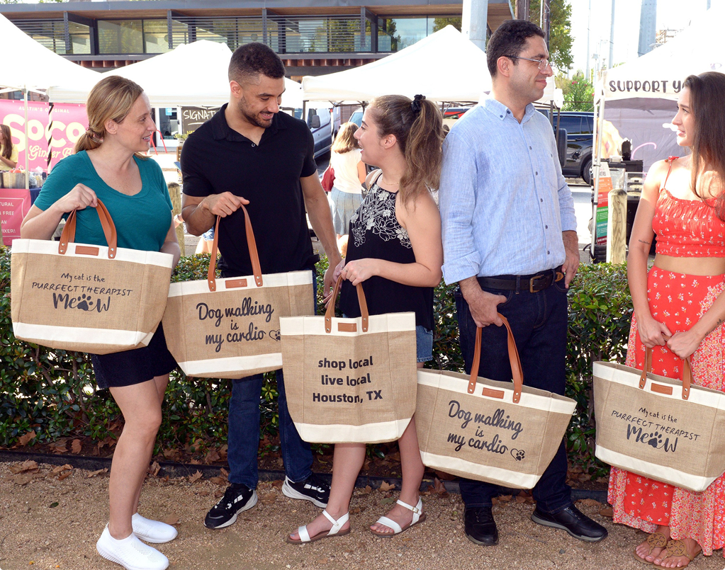 Group of friends outdoors holding Shamajoy’s eco-friendly, reusable bags with fun designs like ‘Dog walking is my cardio’ and ‘My cat is the Perfect therapist’ while shopping locally in Houston, TX. The photo captures the vibrant, sustainable lifestyle promoted by Shamajoy.