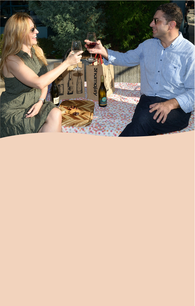 Two people enjoying a wine toast while sitting outdoors with eco-friendly Shamajoy reusable wine bags, surrounded by a bottle of wine and snacks.